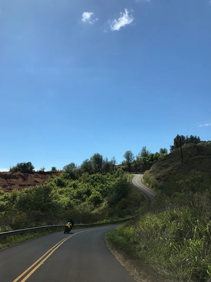 Motorcyclist leaning into a winding mountain road through lush green Hawaiian hills under blue sky