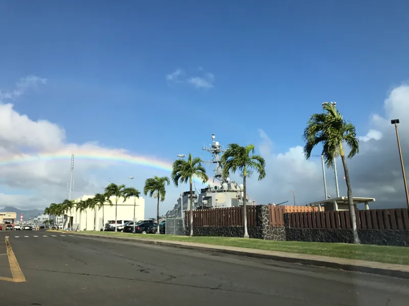 Rainbow arching over palm trees and a Navy ship docked at Pearl Harbor on a partly cloudy day