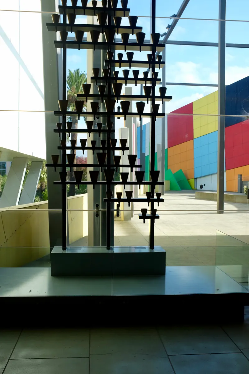 Tall black metal bell sculpture silhouetted against glass walls and a colorful building facade in a museum lobby