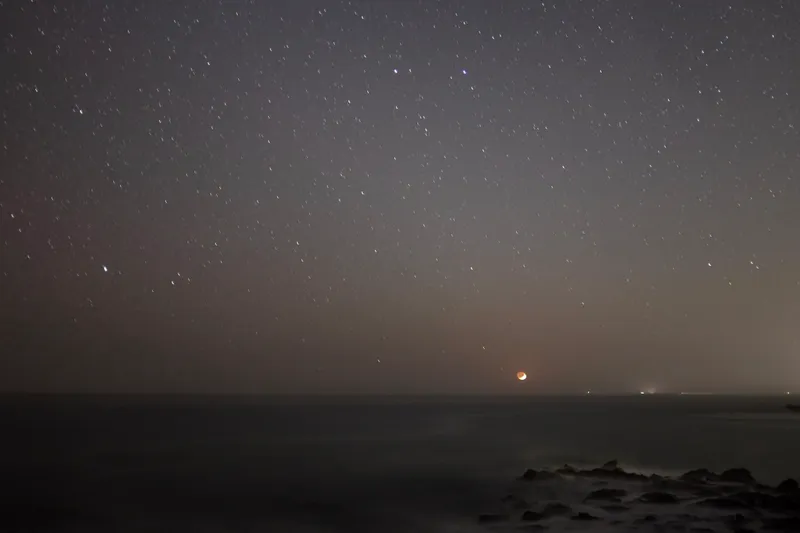Stars scattered across a dark sky over a calm ocean with a faint orange moon near the horizon