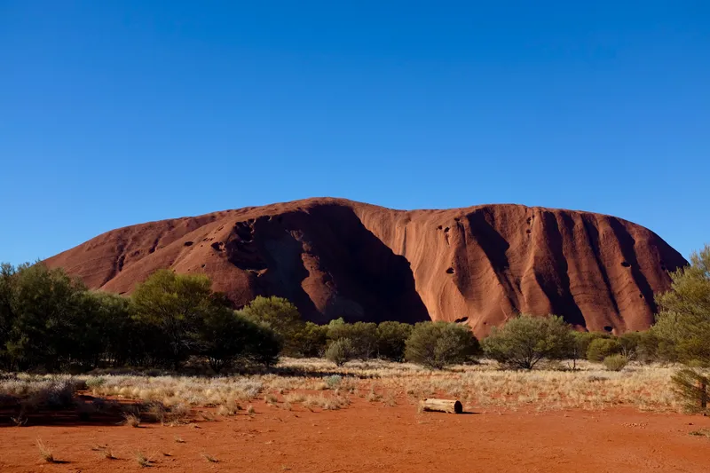 Uluru rising from red desert sand and scrubland under a deep blue cloudless sky