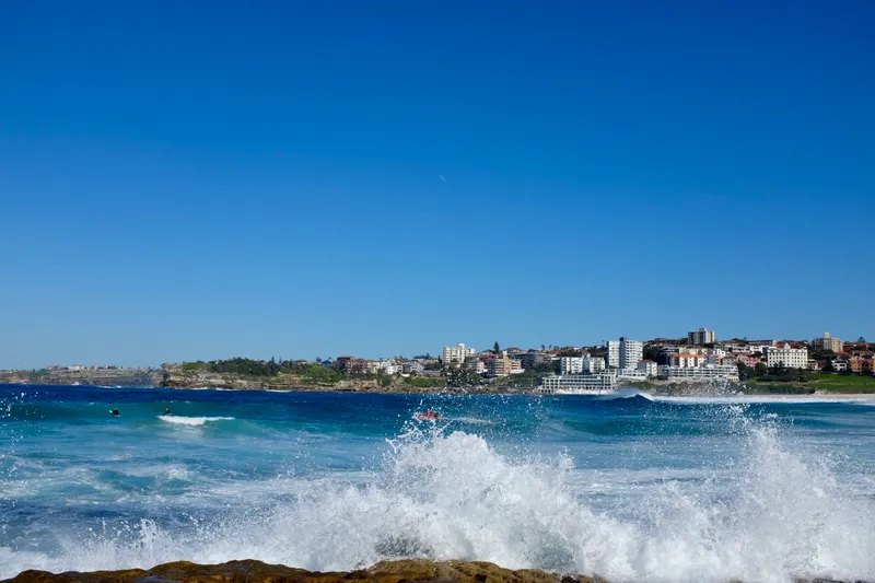 Waves crashing onto rocks at Bondi Beach with the coastal cliffs and buildings beyond under clear blue sky