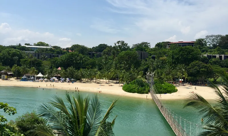 White sand beach and turquoise water at Sentosa Island with a suspension bridge and tropical trees